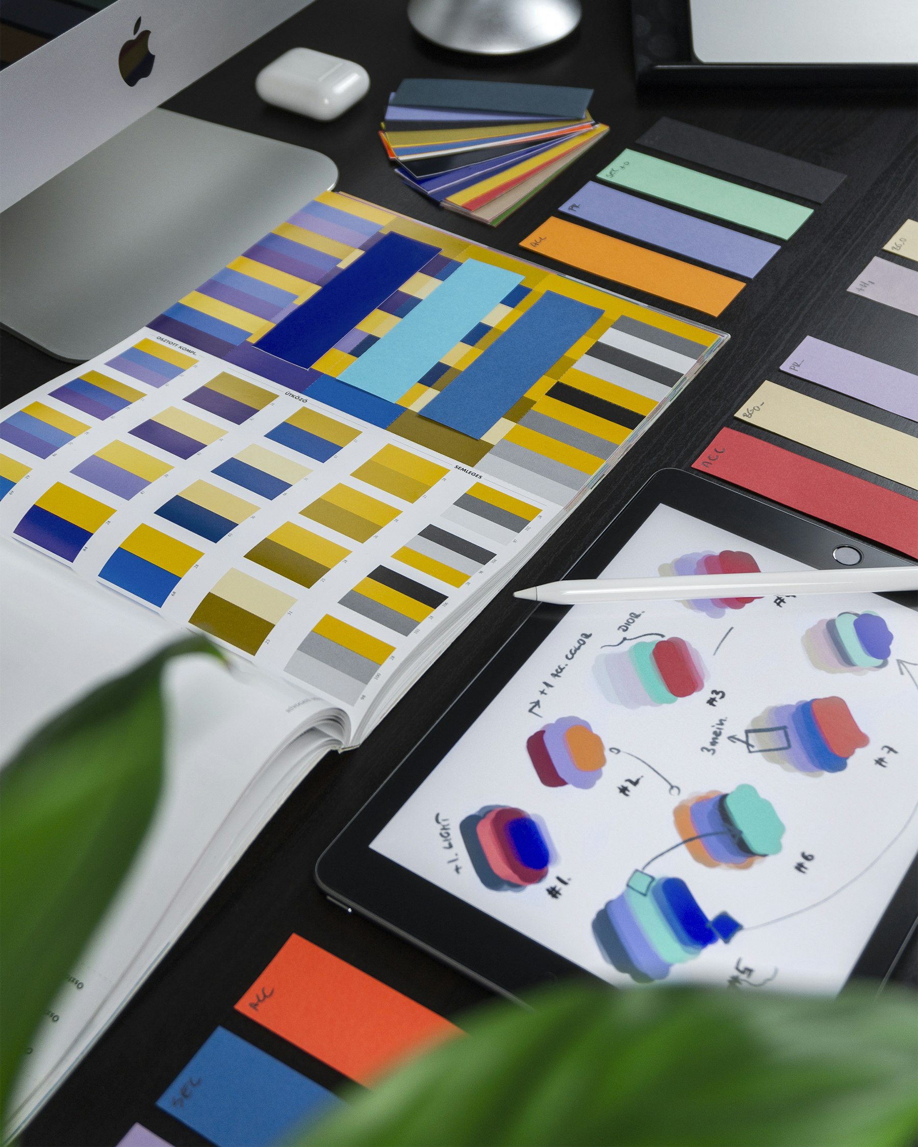 Colour swatches, a colour reference book and a tablet with palette sketches on a dark desk